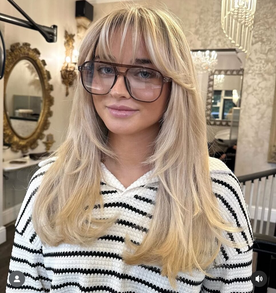 A woman with blonde hair and glasses wearing a striped sweater, featuring light and airy wispy bangs that provide a soft, feathered frame for the face.