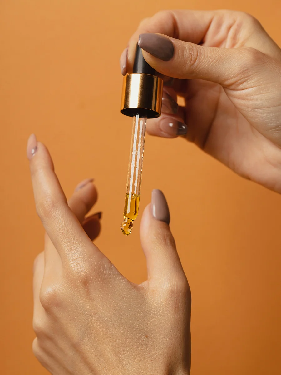 Een close-up van twee gemanicuurde handen, waarvan één hand een glazen pipet vasthoudt waar nagelriemolie uit druppelt