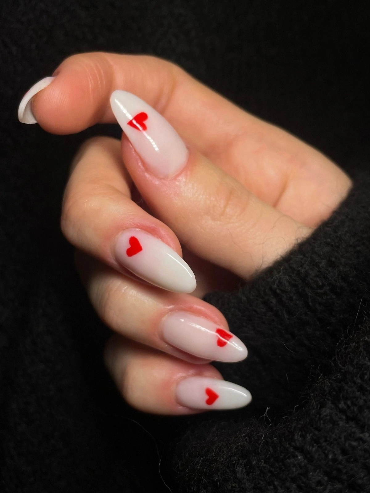 Close-up van de hand van een vrouw met witte acrylnagels in amandelvorm, versierd met kleine rode hartjes