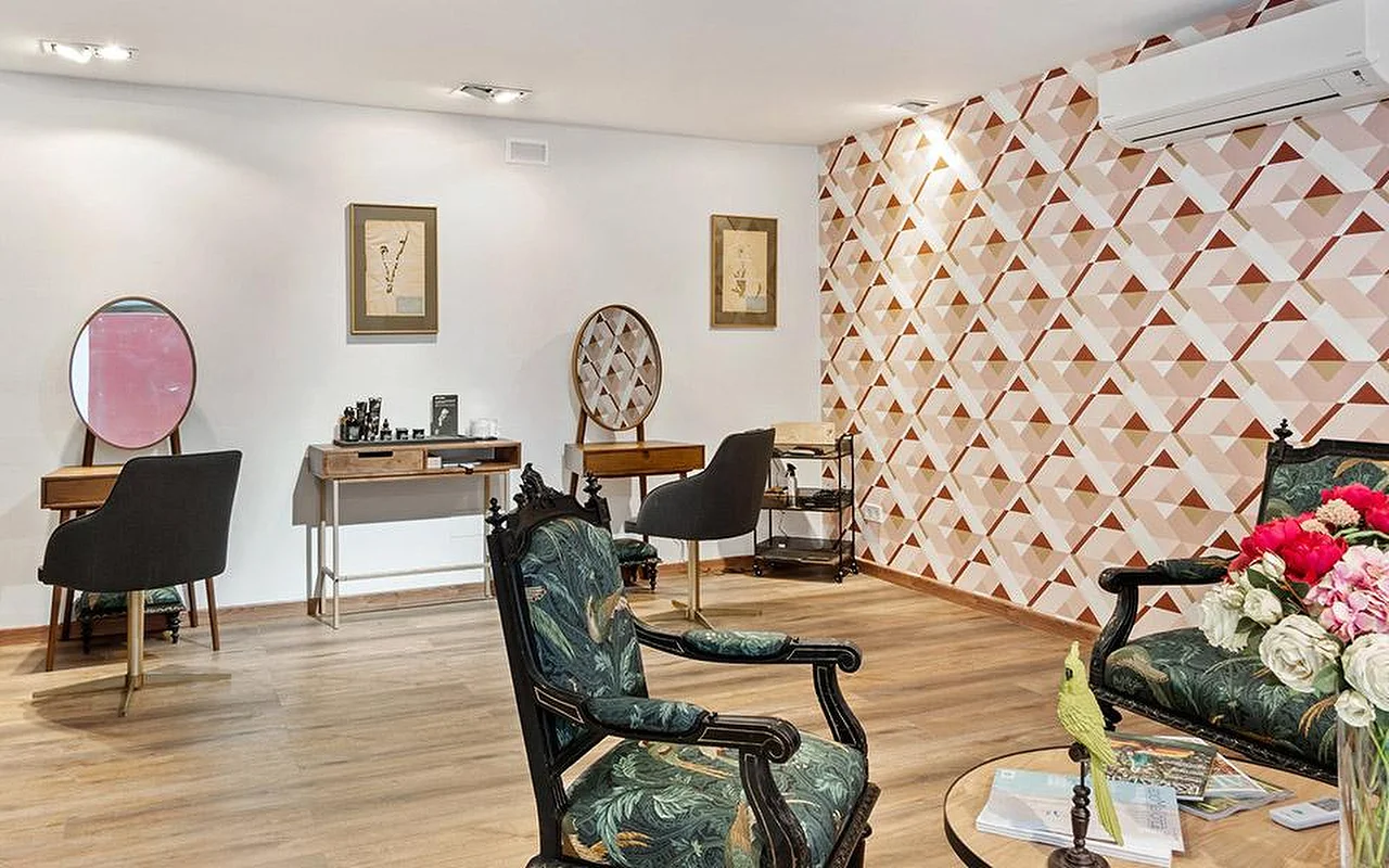 Een elegante salonruimte met een geometrische roze-terracotta accentmuur, een paar sierlijk gesneden stoelen bekleed met groene botanische stof, houten kaptafels met ronde spiegels en op de voorgrond een vaas met roze en rode bloemen.