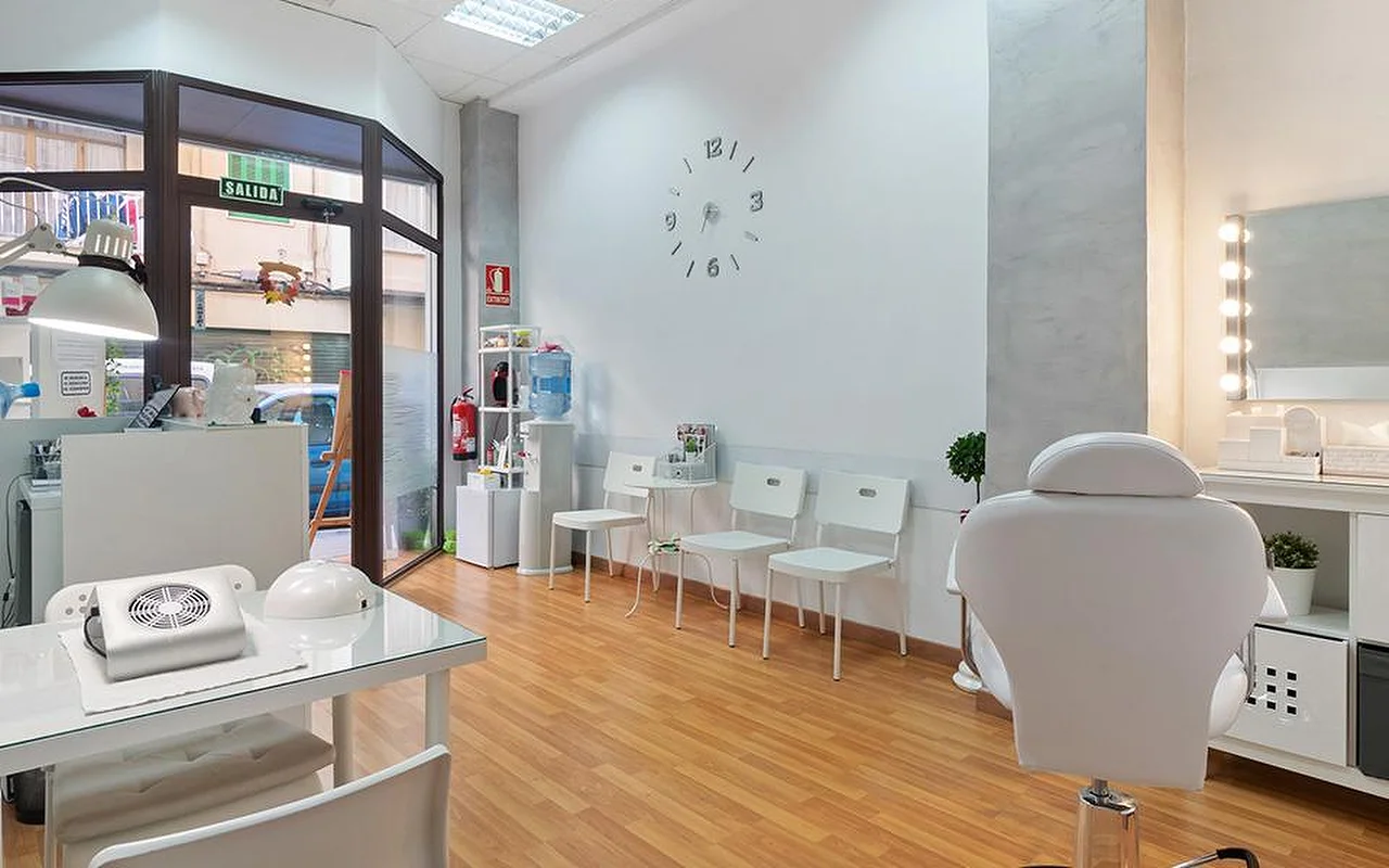 Een lichte, witte salon met lichte houten vloeren, een rij witte wachtstoelen, een minimalistische wandklok, een verlichte make-upspiegel, een witte verstelbare behandelstoel en een manicuretafel bij het raam.