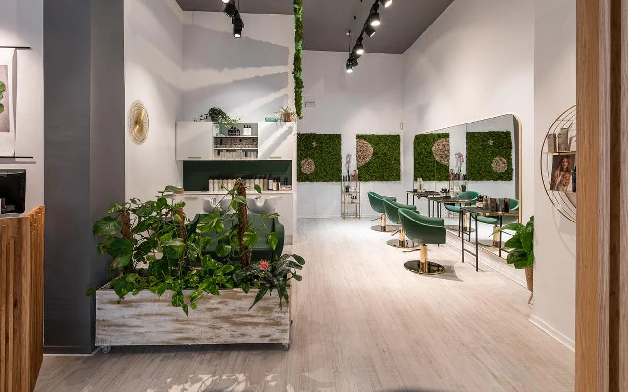 Een kapsalon vol planten met lichte houten vloeren, een verweerde witte plantenbak die overloopt van het groen, mos-wandkunst, smaragdgroene velours kappersstoelen met gouden details en een wasstation erachter.