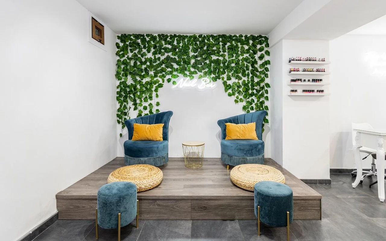 Een hoek van een nagelsalon met een cascade van groene planten aan de muur en een klein neonbordje, twee fauteuils van turquoise velours met mosterdgele kussens op een verhoogd houten platform, gevlochten poefjes en rechts schappen vol kleurrijke nagellak.
