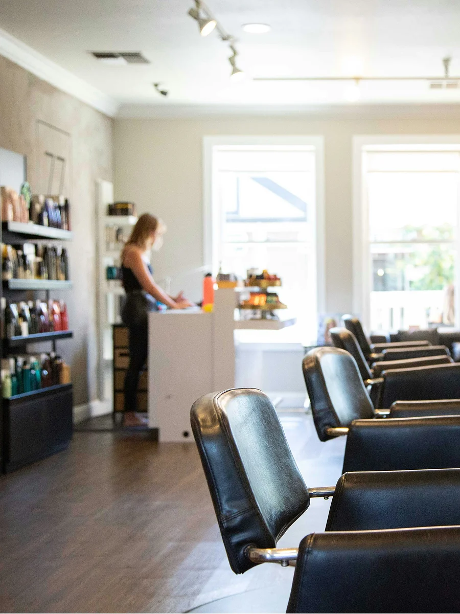 Interieurfoto van een schone, lichte en goed geventileerde kapsalon met grote ramen, zwarte stoelen en schappen vol producten