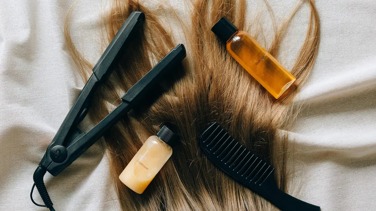 Flatlay van haarverzorgingsproducten op iemands lange steile haar, met shampoo, conditioner, een stijltang en een grove kam