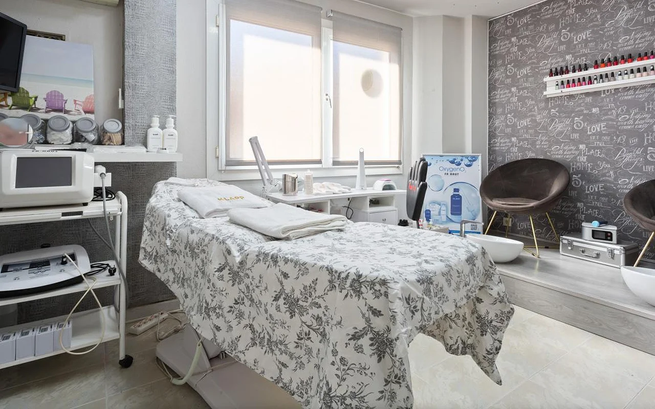 Een lichte behandelkamer met een bed met grijs gebloemd linnen en opgevouwen witte handdoeken, aesthetics-apparaten op trolleys links, een raam met de jaloezie half neer, en een verhoogd gedeelte met een bruine velours stoel en pedicurebakjes tegen een accentmuur met handschrift-print.