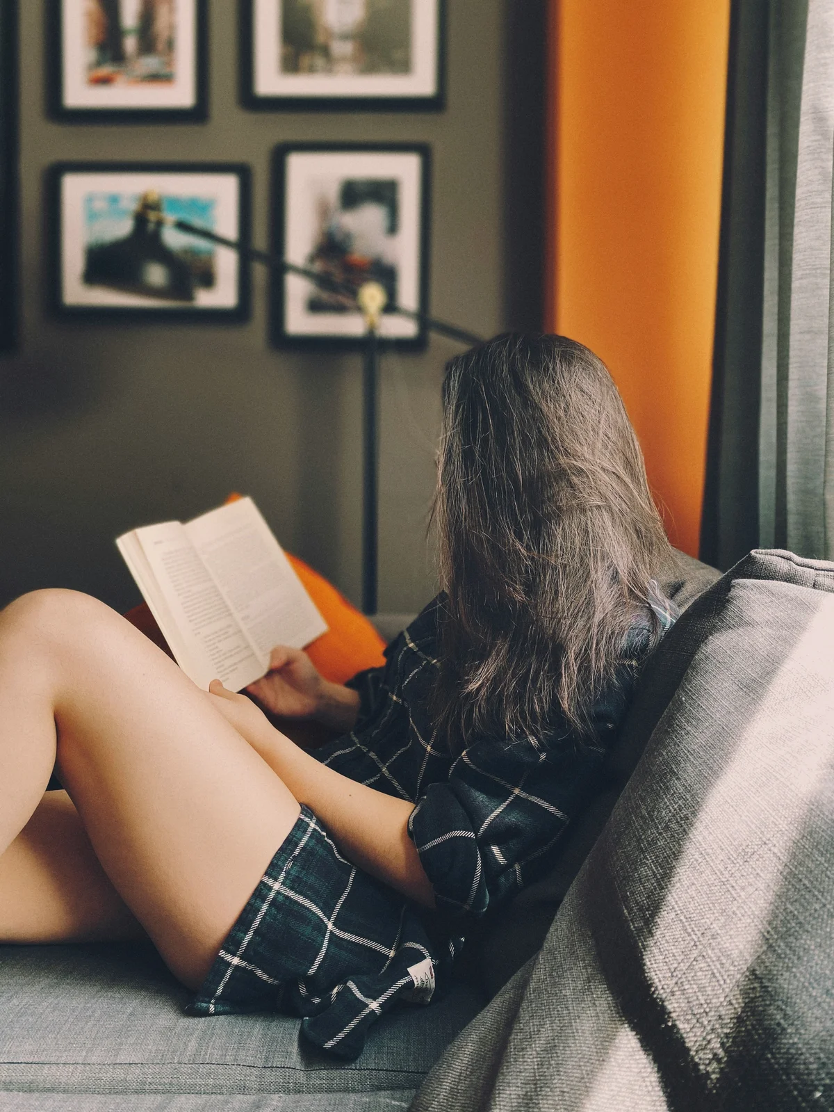 Zijaanzicht van een vrouw die op een bank zit te lezen, haar lange haar bedekt haar gezicht; ze draagt een ruimzittende katoenen short en een donkergroen geruit overhemd
