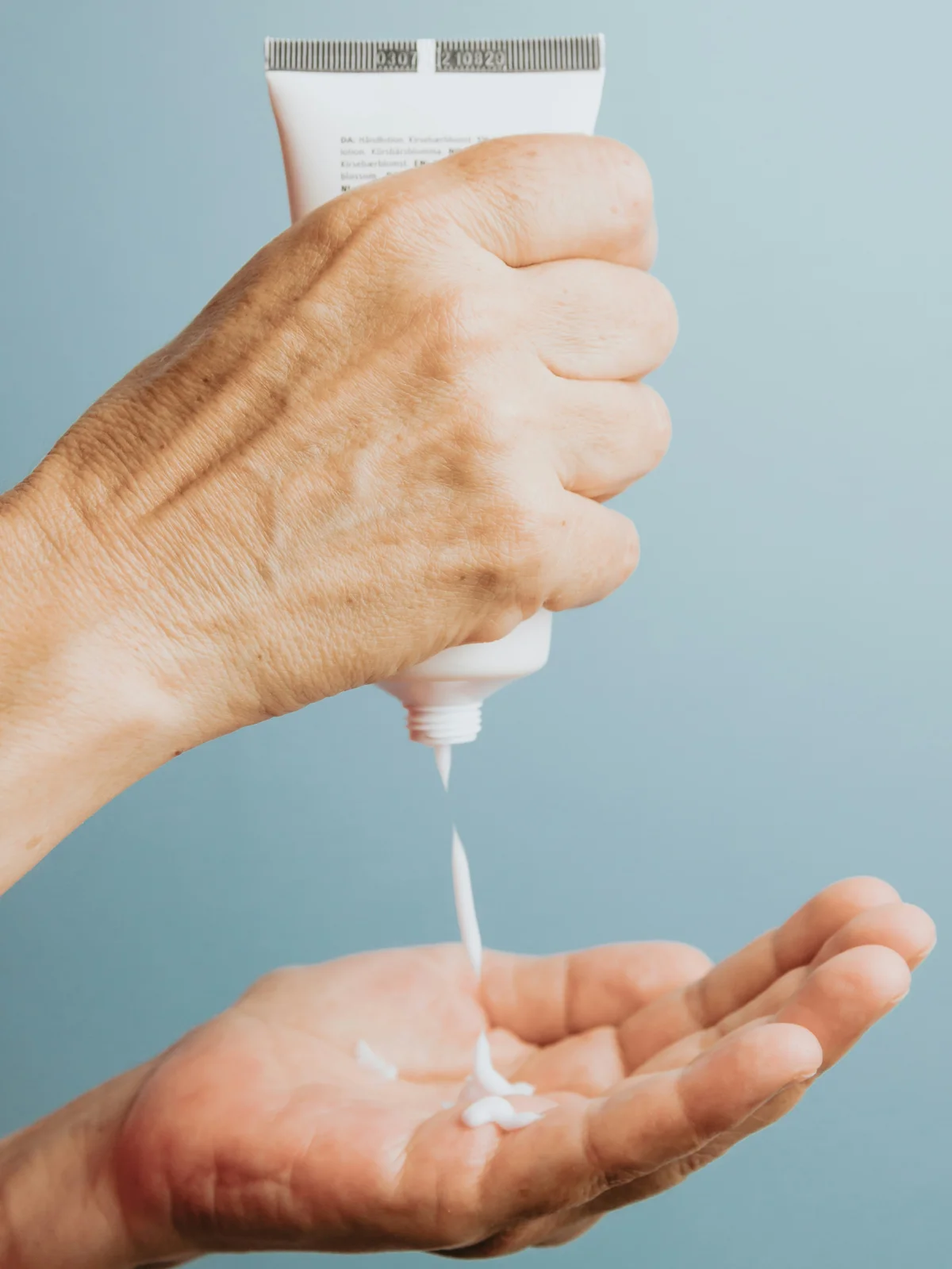 Close-up van een hand die een tube witte bodylotion rechtop vasthoudt en er een beetje van in de andere hand eronder laat vallen
