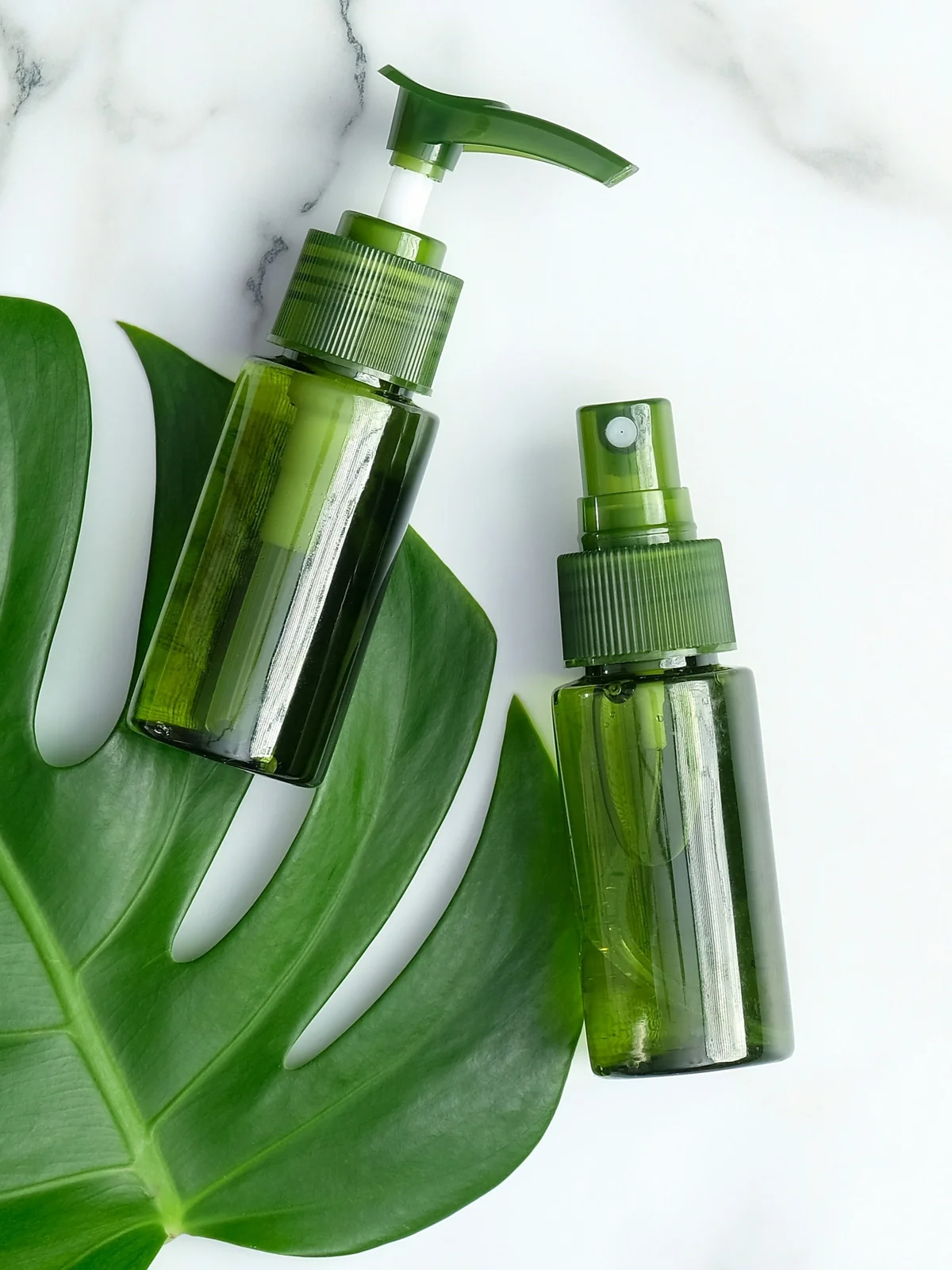 Plat uitgestald: twee groene flesjes met verfrissende gels en serums op een groen blad en een witte marmeren ondergrond