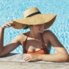 Een vrouw in een zonnig buitenzwembad die tegen de rand leunt, met een oranje bikini en een strohoed met brede rand