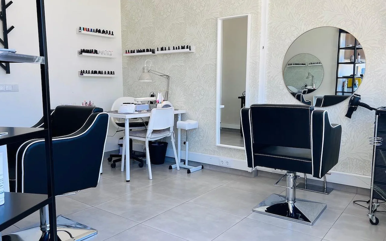 Een compacte haar- en nagelsalon met zwarte, wit afgebiesde stylingstoelen, een ronde spiegel en een hoge spiegel tegen grijs bloemenbehang, en wandplanken vol nagellak.