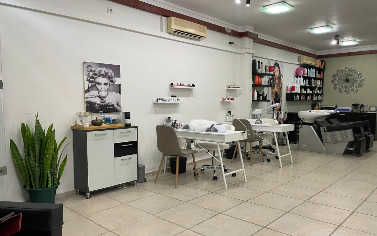 Een haar- en beautysalon met twee witte manicuretafels en taupe stoelen, een wasbak en productplanken langs de muur, een hoge vrouwentong in de hoek en een grote zwart-witfoto van een vrouw met krulspelden.