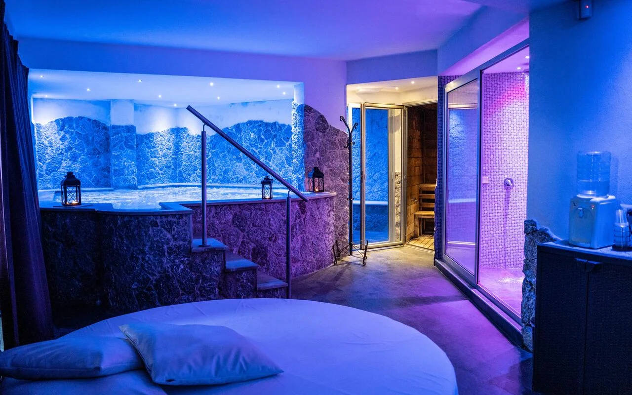 Een privé-spasuite bij Bellezza e Benessere in Taormina, verlicht in blauw en paars, met een verzonken bubbelbad in een stenen ombouw, een houten sauna en daarachter een met mozaïek betegeld stoombad, en een rond bed op de voorgrond.