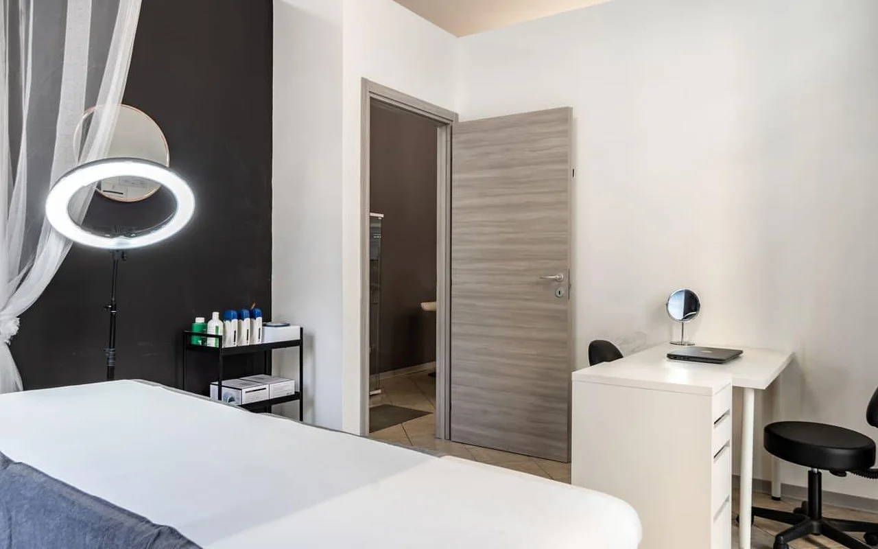 Een behandelcabine bij Coquette Wellness and Beauty, met een witte behandeltafel, een verlicht ringlicht op een statief tegen een zwarte accentmuur, lichte gordijnen voor het raam en een klein wit bureau met kaptafelspiegel naast een deur naar een eigen badkamer.