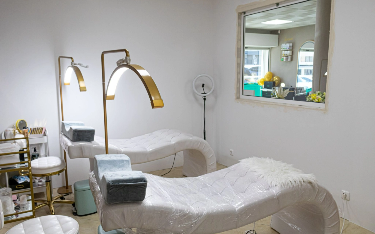 Een lichte beautybehandelruimte met twee witte gewatteerde ligstoelen, hoge gouden booglampen en een ringlamp, met een intern raam dat uitkijkt op de receptie.