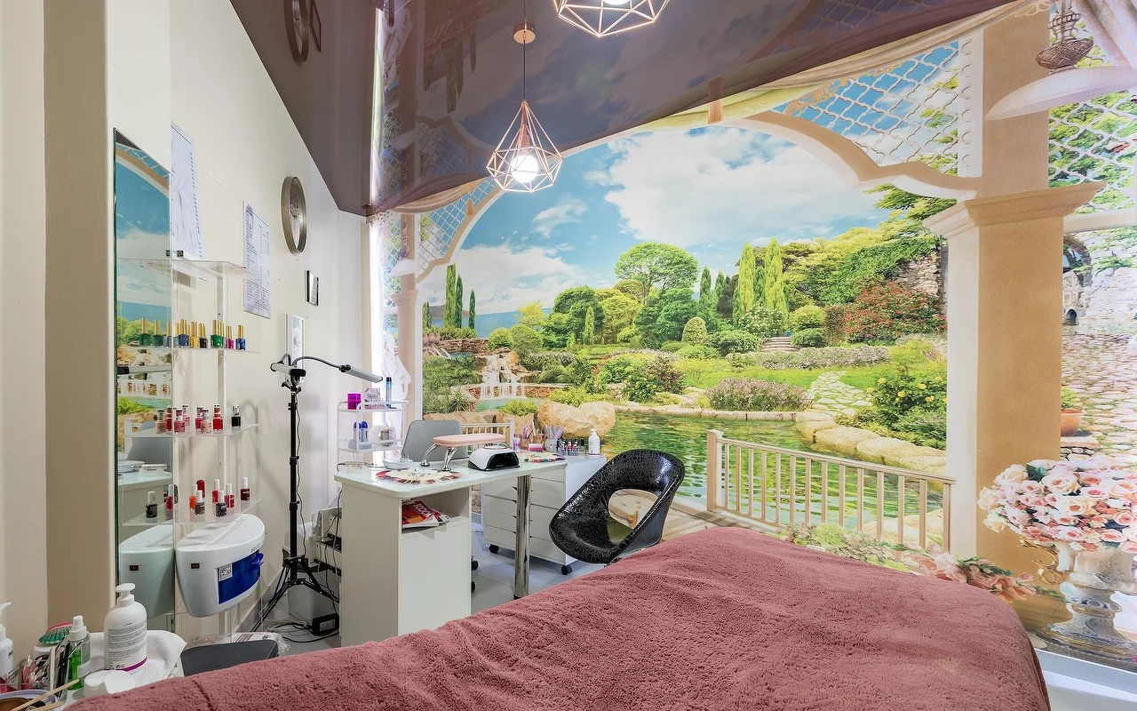 Een beautybehandelruimte met een roze gedrapeerd bed en een witte nagelstation, tegen een grote muurschildering met tuin en meer, met glazen planken vol producten ernaast.