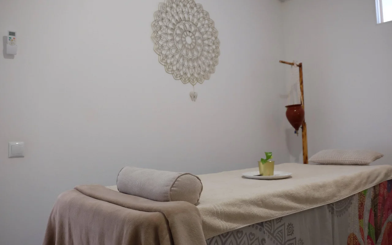 Een rustige massageruimte in zachte, neutrale tinten, met een massagetafel met handdoek en een nekrol, een grote gehaakte mandala aan de muur en een houten standaard met een hangende terracottapot.
