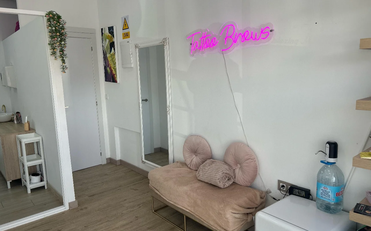 Een behandelplek bij YG Beauty Estudio in Torremolinos, met een roze 'Tattoo Brows'-neonbord, een passpiegel en een gestoffeerde beige ligbank.